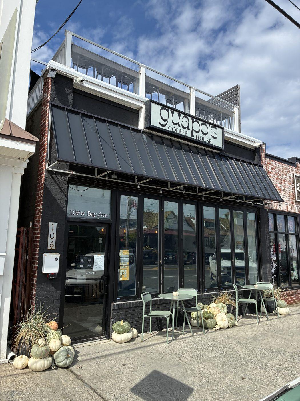 A dark storefront with a long run of white and pale green pumpkins along the sidewalk, the doorway and the café tables.